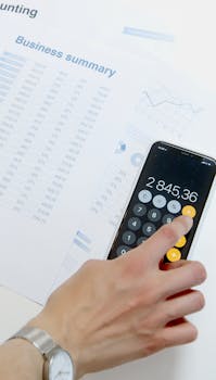 Hand using smartphone calculator for analyzing business summary and accounting data.