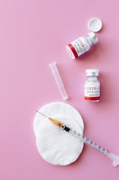 Healthcare flatlay featuring COVID-19 vaccine vials and syringe on a pink surface.