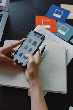 A woman browsing fashion items on a smartphone while sitting at a desk indoors.